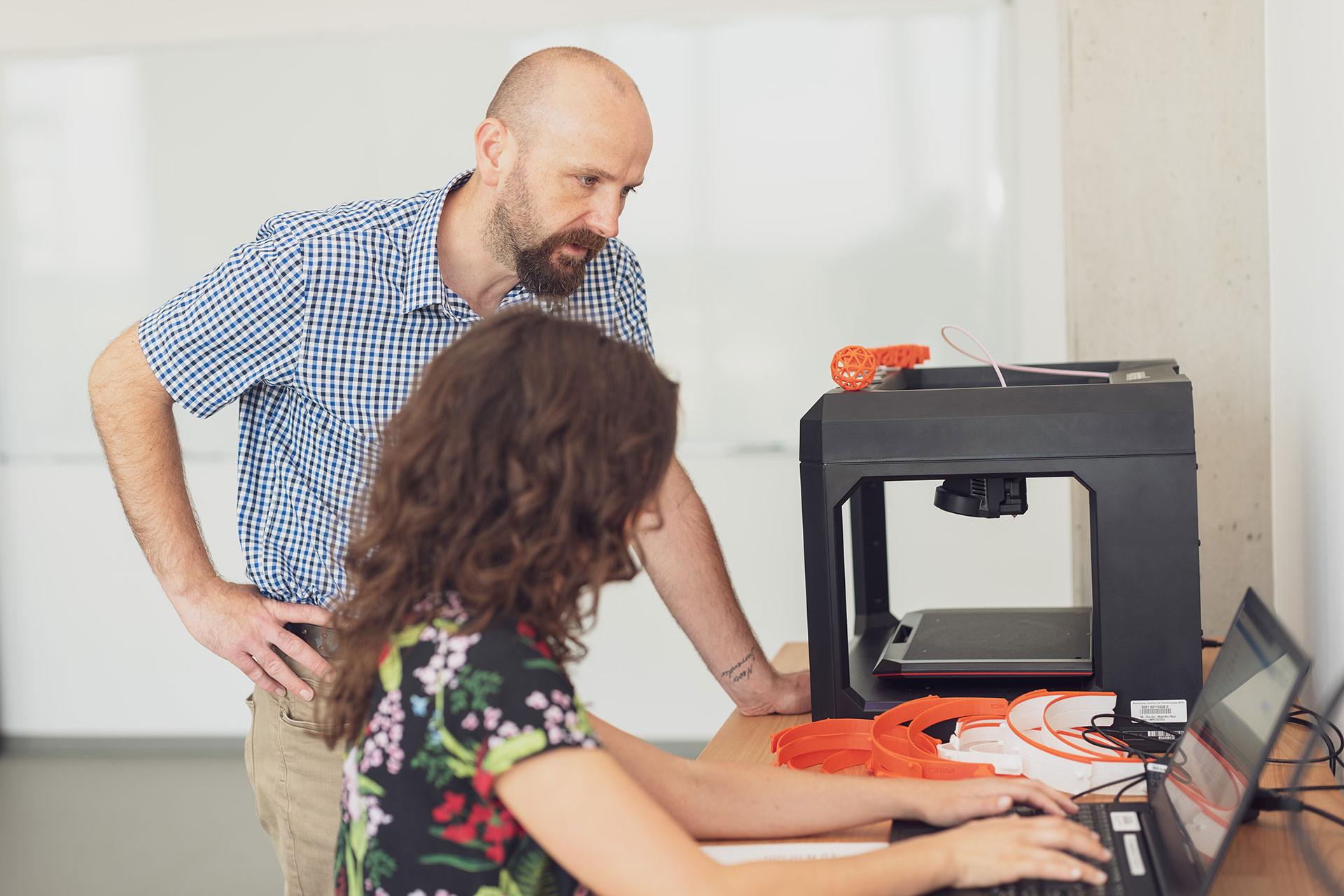 Zwei Personen arbeiten im Makerspace an einem Schreibtisch mit einem Laptop und einem 3D-Drucker.