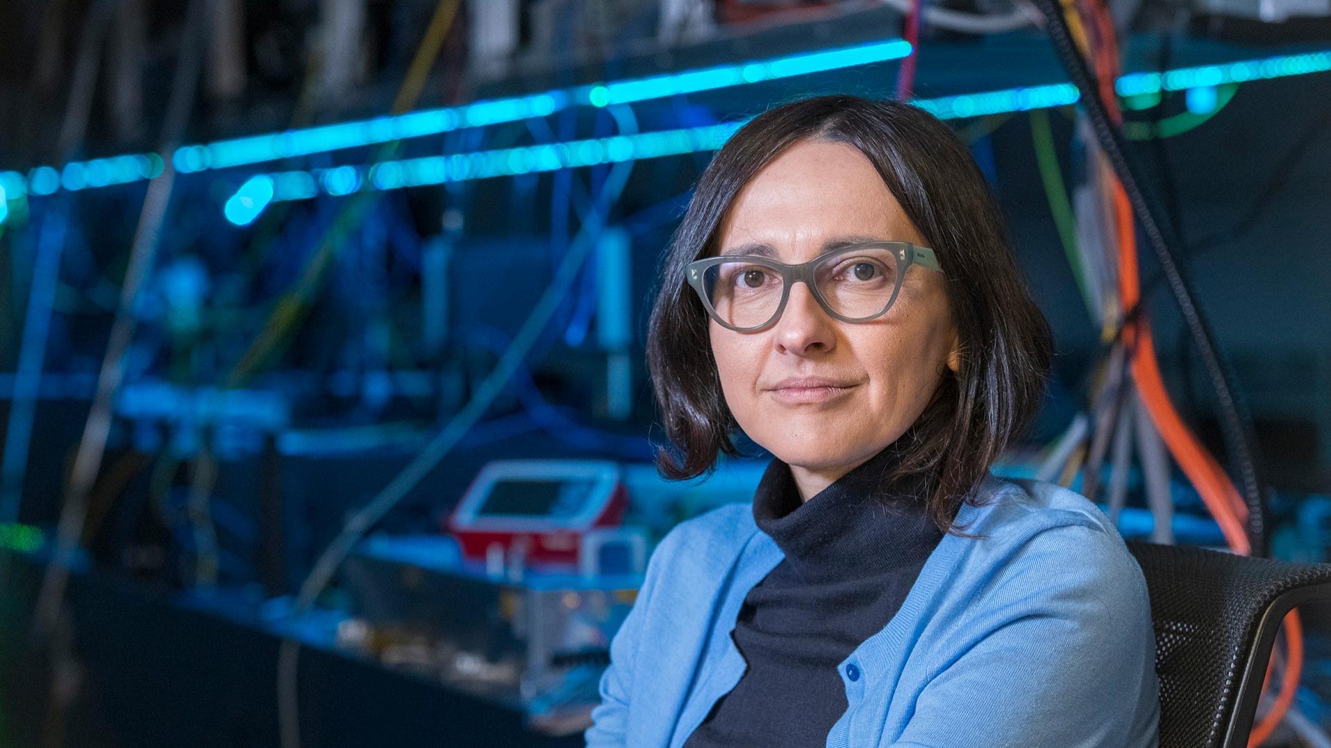 Professorin Dr. Jelena Vuckovic sitzt in einem Labor mit technischen Geräten im Hintergrund. Sie trägt einen schwarzen Rollkragenpullover und eine hellblaue Strickjacke.
