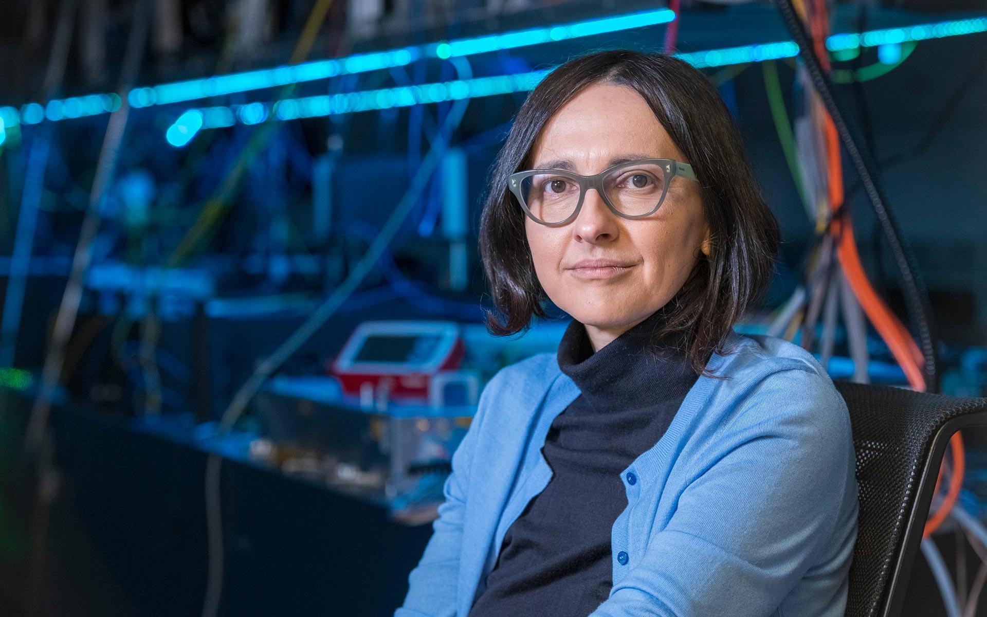 Professorin Dr. Jelena Vuckovic sitzt in einem Labor mit technischen Geräten im Hintergrund. Sie trägt einen schwarzen Rollkragenpullover und eine hellblaue Strickjacke.