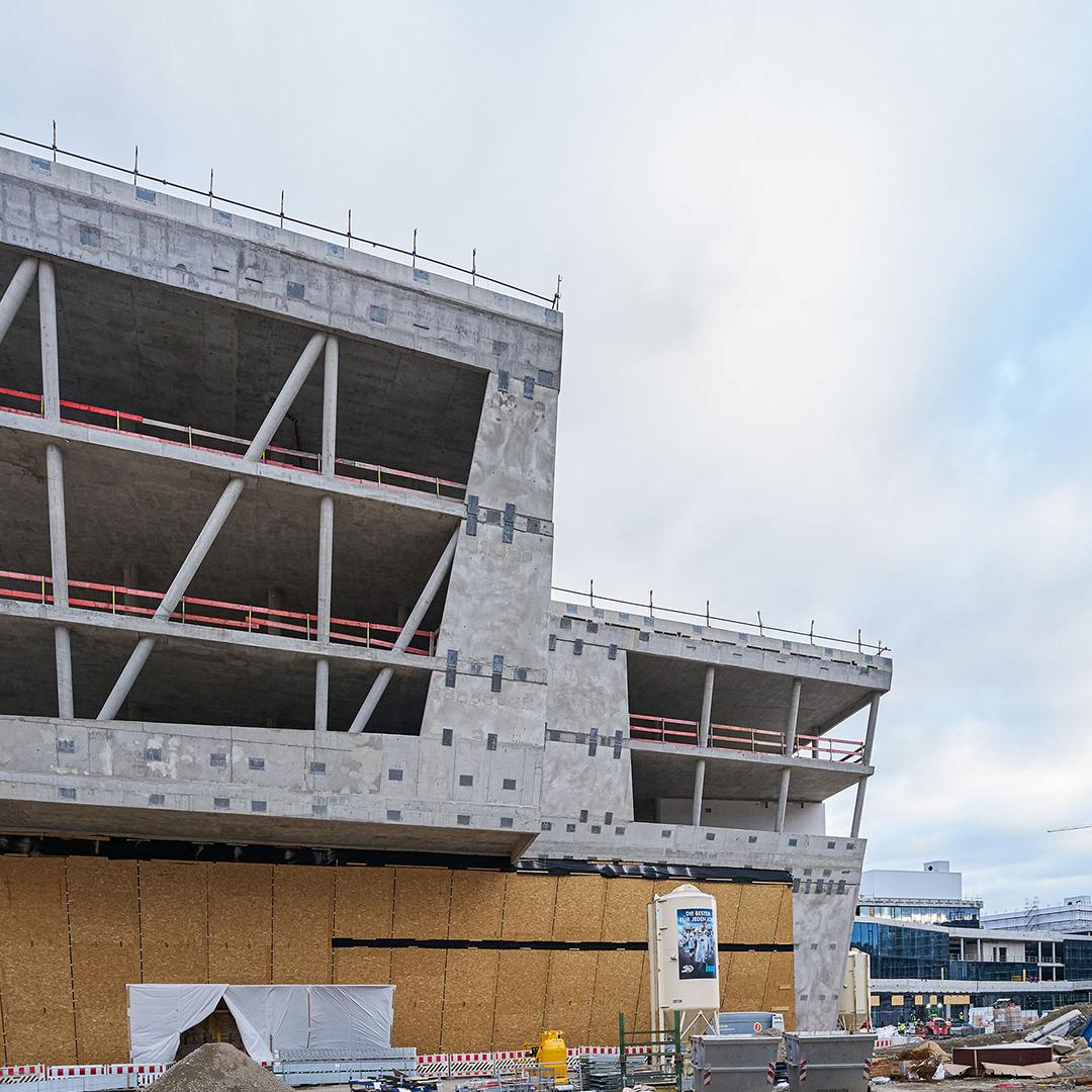 Unfertiges, modernes Gebäude mit mehreren überhängenden Stockwerken und sichtbarer Stahlbetonstruktur unter bewölktem Himmel.