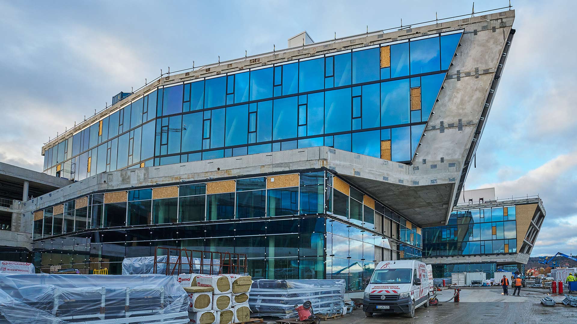 Modernes, mehrstöckiges Gebäude mit blauen Glasfassaden und sichtbaren Baustellenmaterialien davor, mehrere Bauarbeiter und ein weißer Transporter stehen auf der Baustelle.