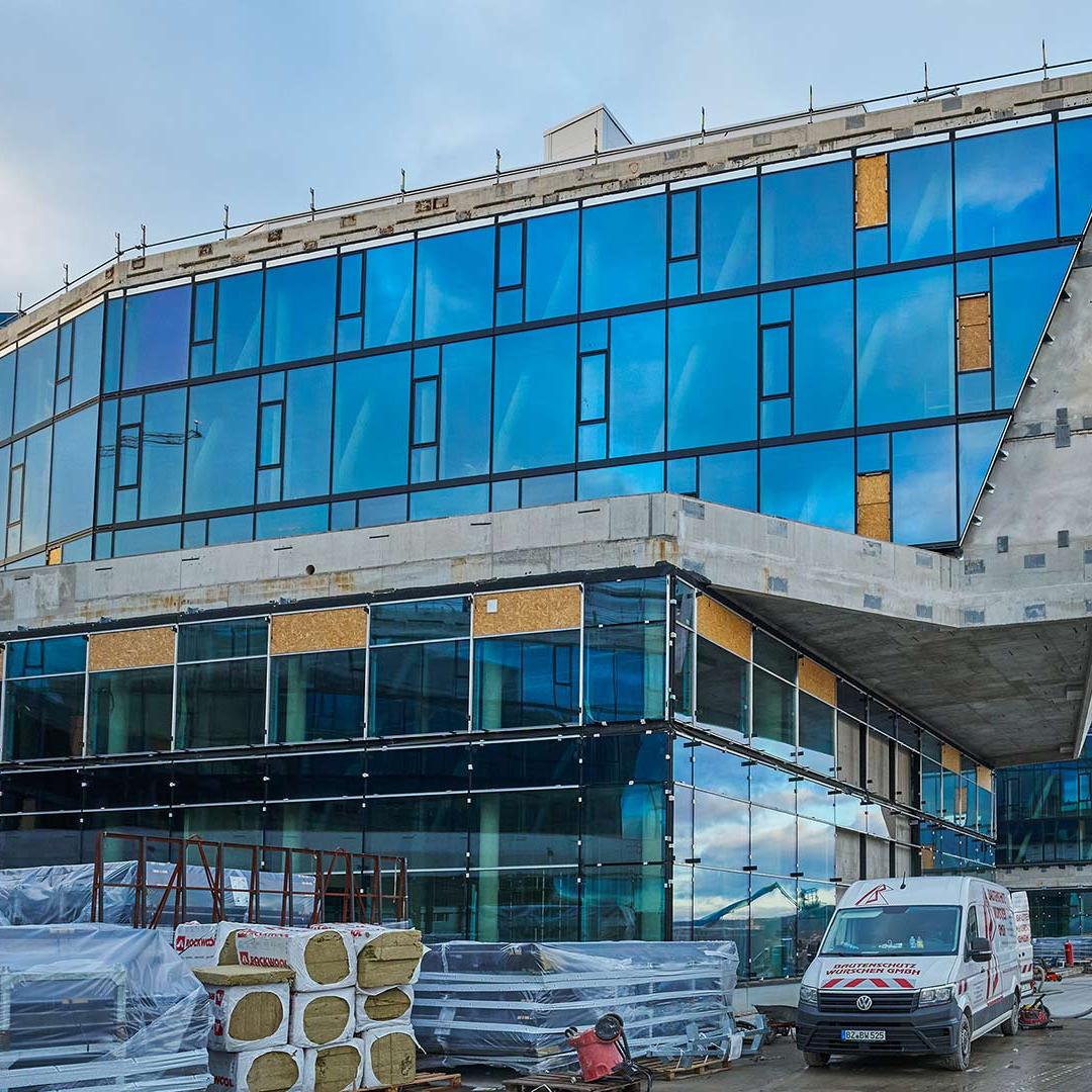 Modernes, mehrstöckiges Gebäude mit blauen Glasfassaden und sichtbarer Baustelle im Vordergrund, mehrere Fahrzeuge und Bauarbeiter erkennbar.