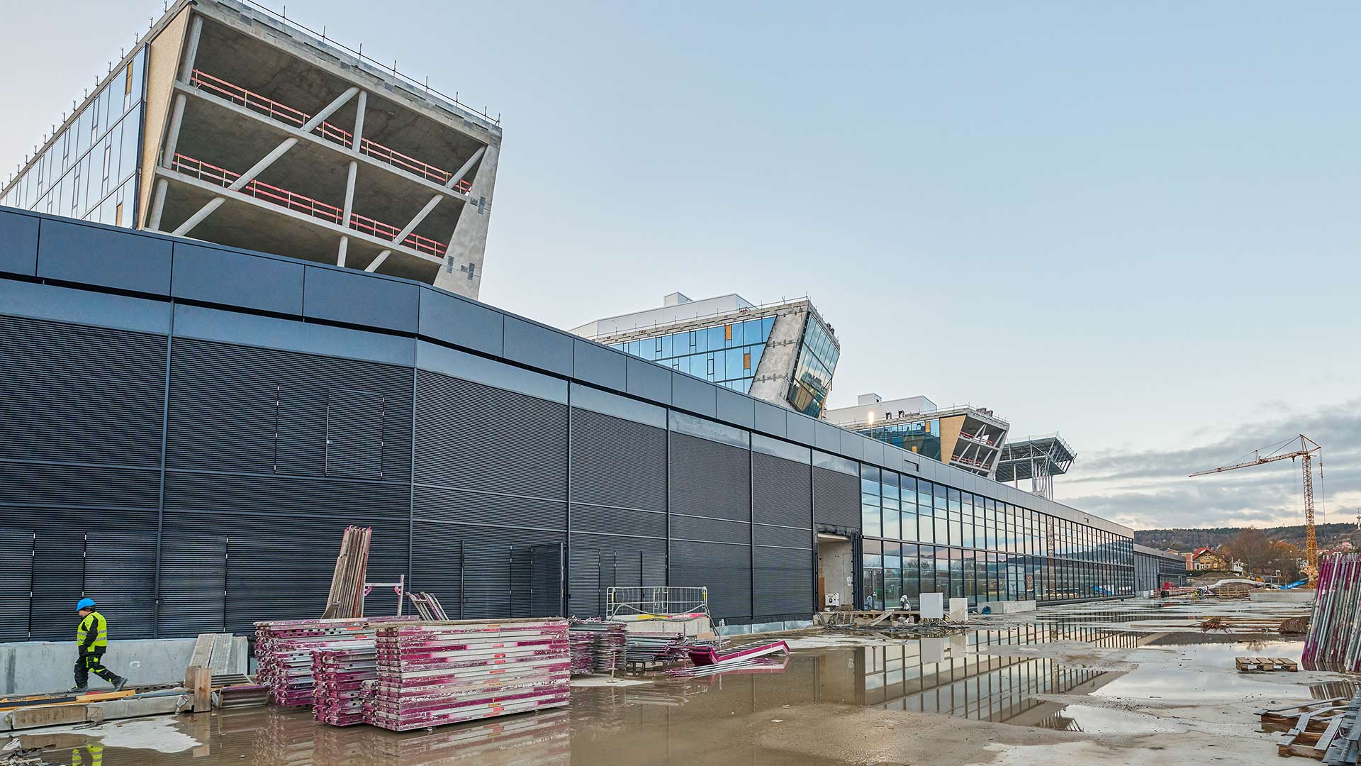 Große Baustelle mit langem, dunklem Gebäude, mehreren gestapelten Baumaterialien und Baukran im Hintergrund bei bewölktem Himmel.