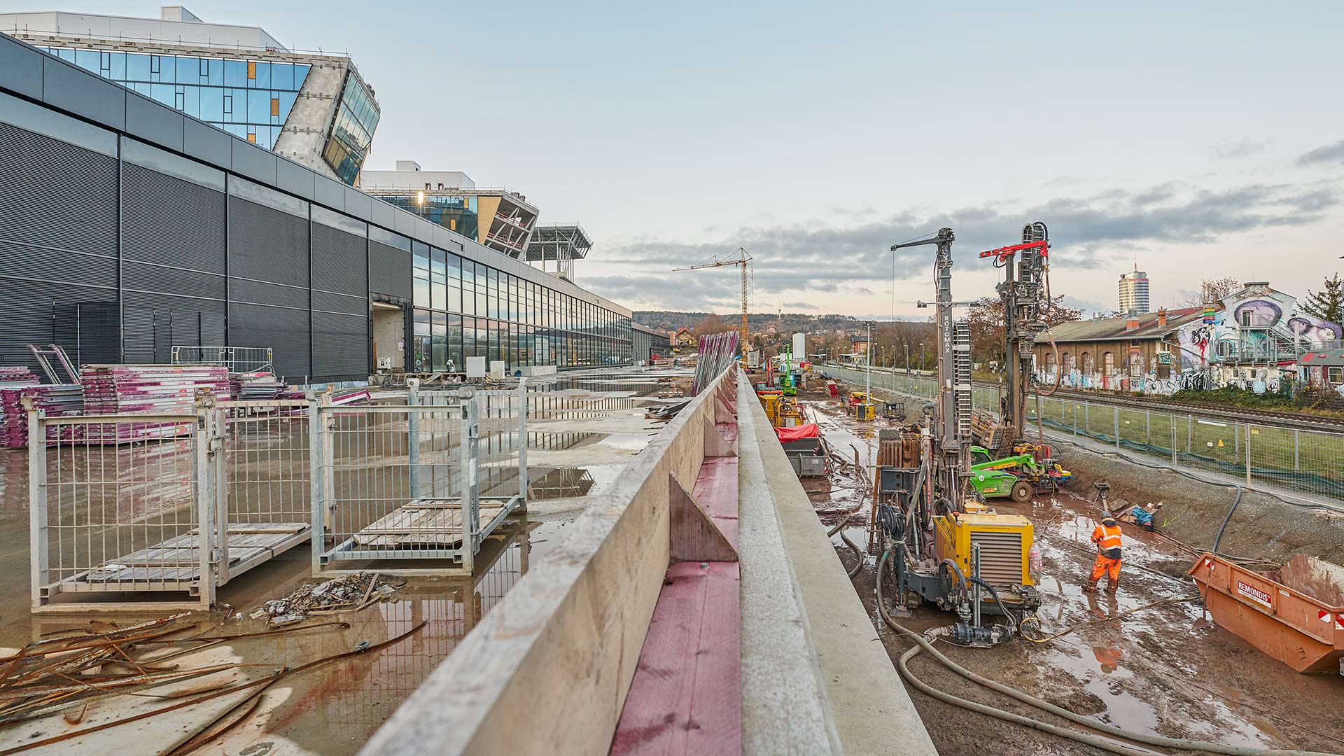 Bauarbeiter in orangener Schutzkleidung arbeiten auf einer Baustelle neben einem Gleis mit Baugeräten und Containern, im Hintergrund Gebäude und Kräne.