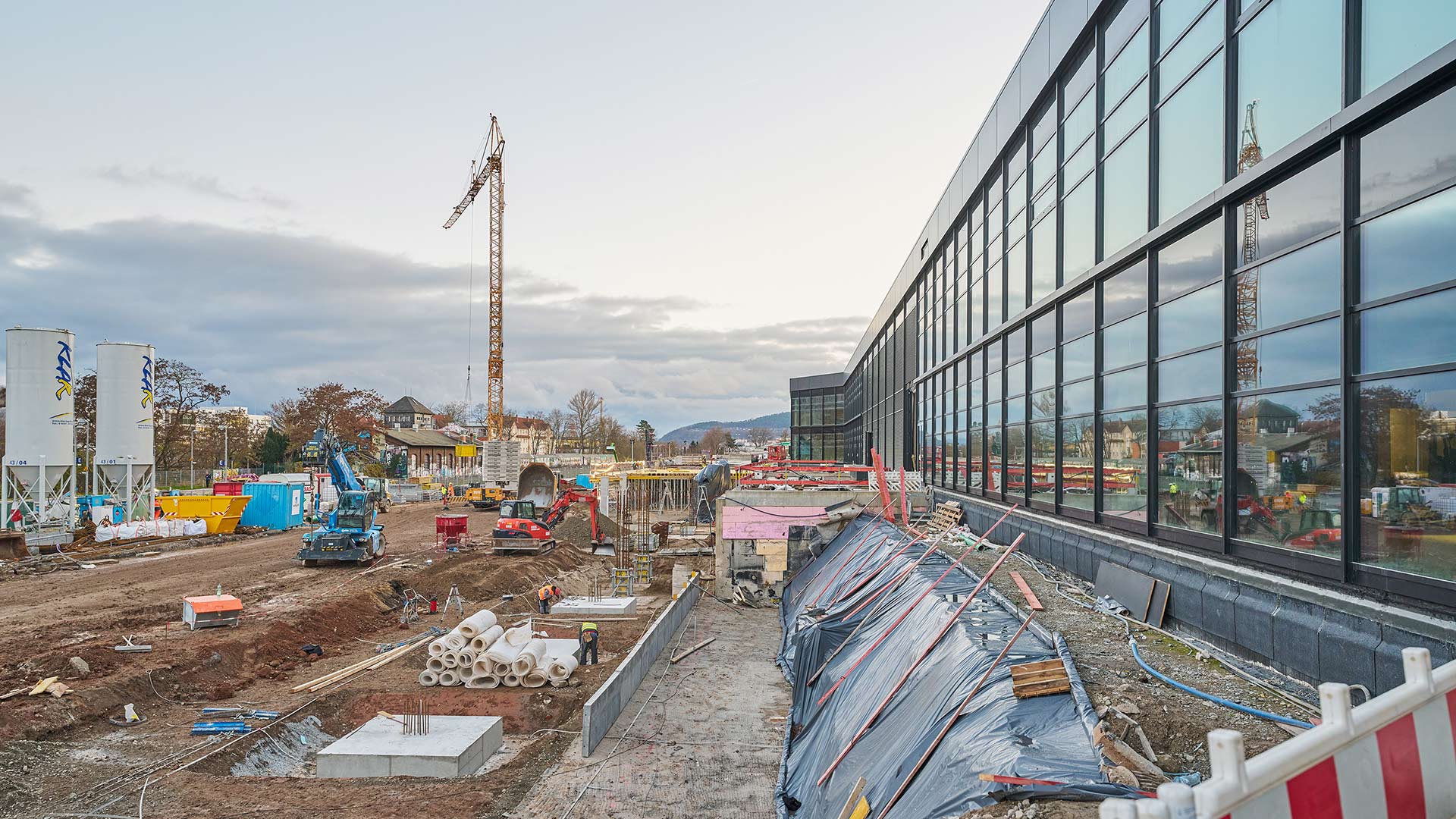 Baustelle mit mehreren Baufahrzeugen, Betonrohren und einem Kran neben einem modernen Glasgebäude unter bewölktem Himmel.