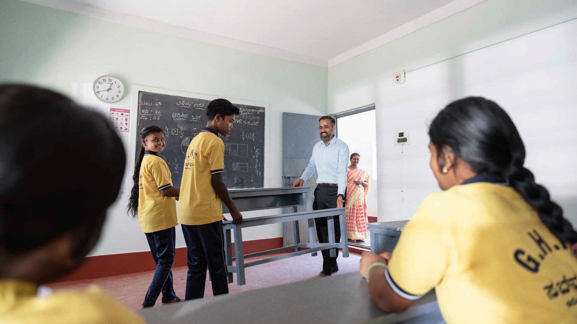 Schüler, Schülerinnen und Manoj Sharma tragen neue Schulbänke in einen Klassenraum