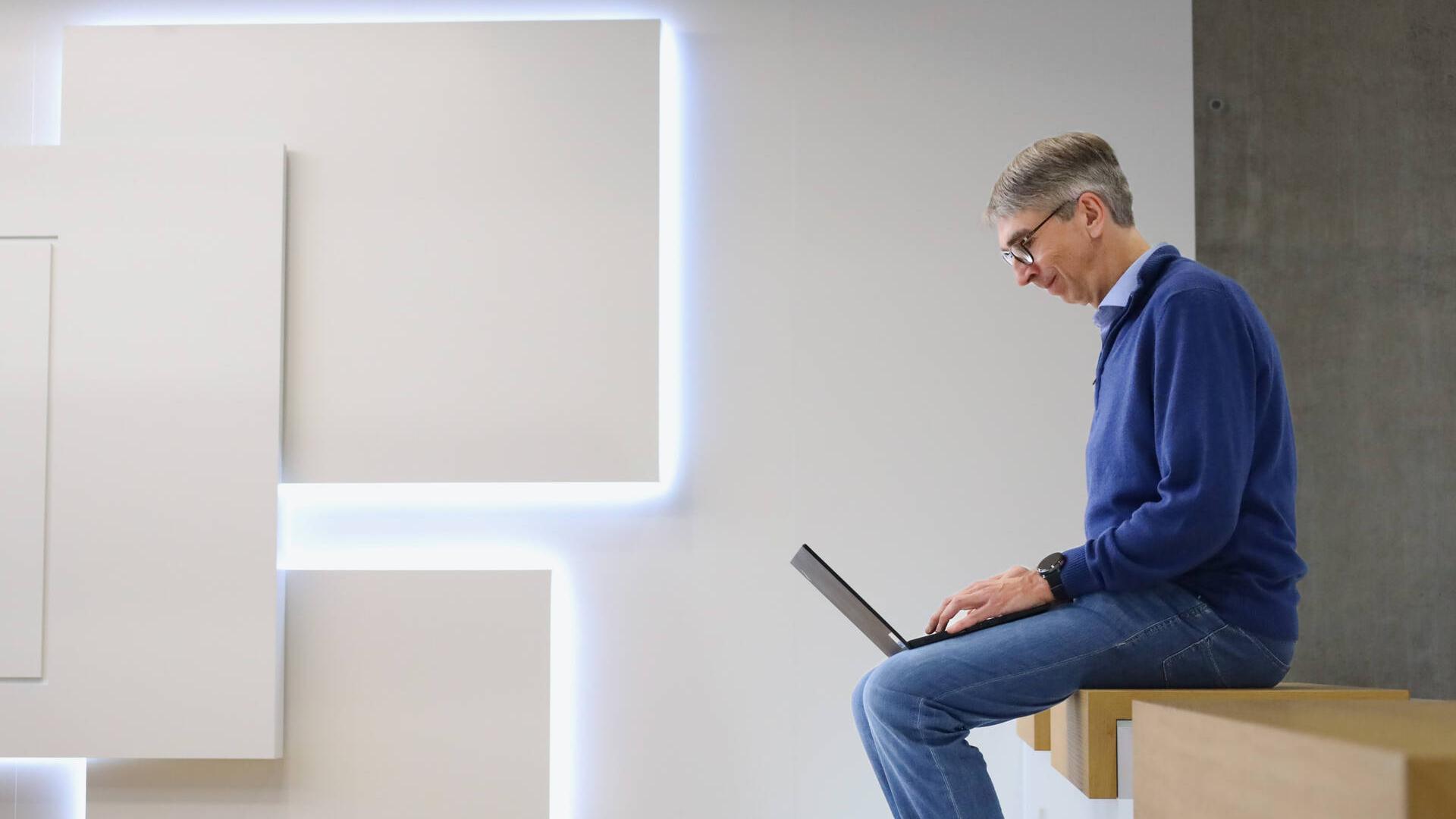 Dr. Ulf Wilhelm sitzt auf einer Holzbank in einem Gemeinschaftsraum bei der Arbeit.