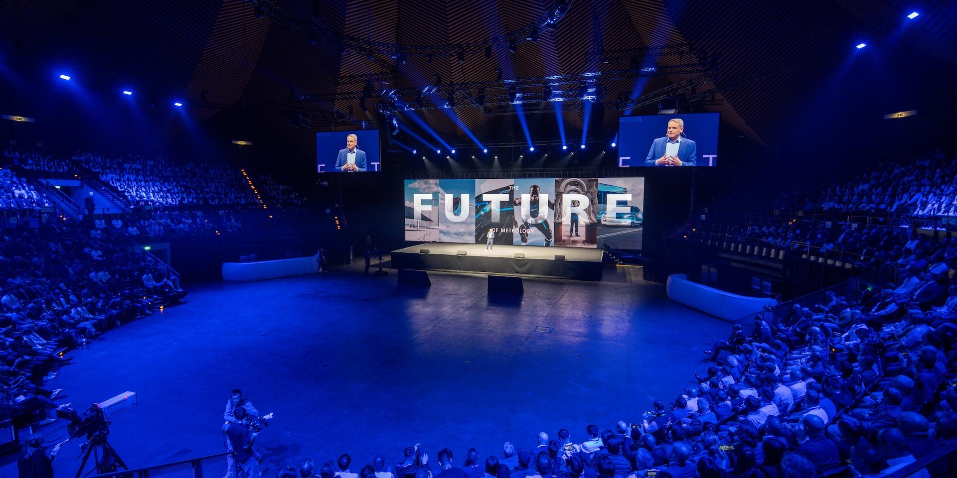 Ein großes Publikum verfolgt eine ZEISS Veranstaltung in einem kreisförmigen Auditorium mit einer prominenten Bühne, auf der das Wort „FUTURE“ steht, sowie Lautsprecher unter dramatischer blauer Beleuchtung.
