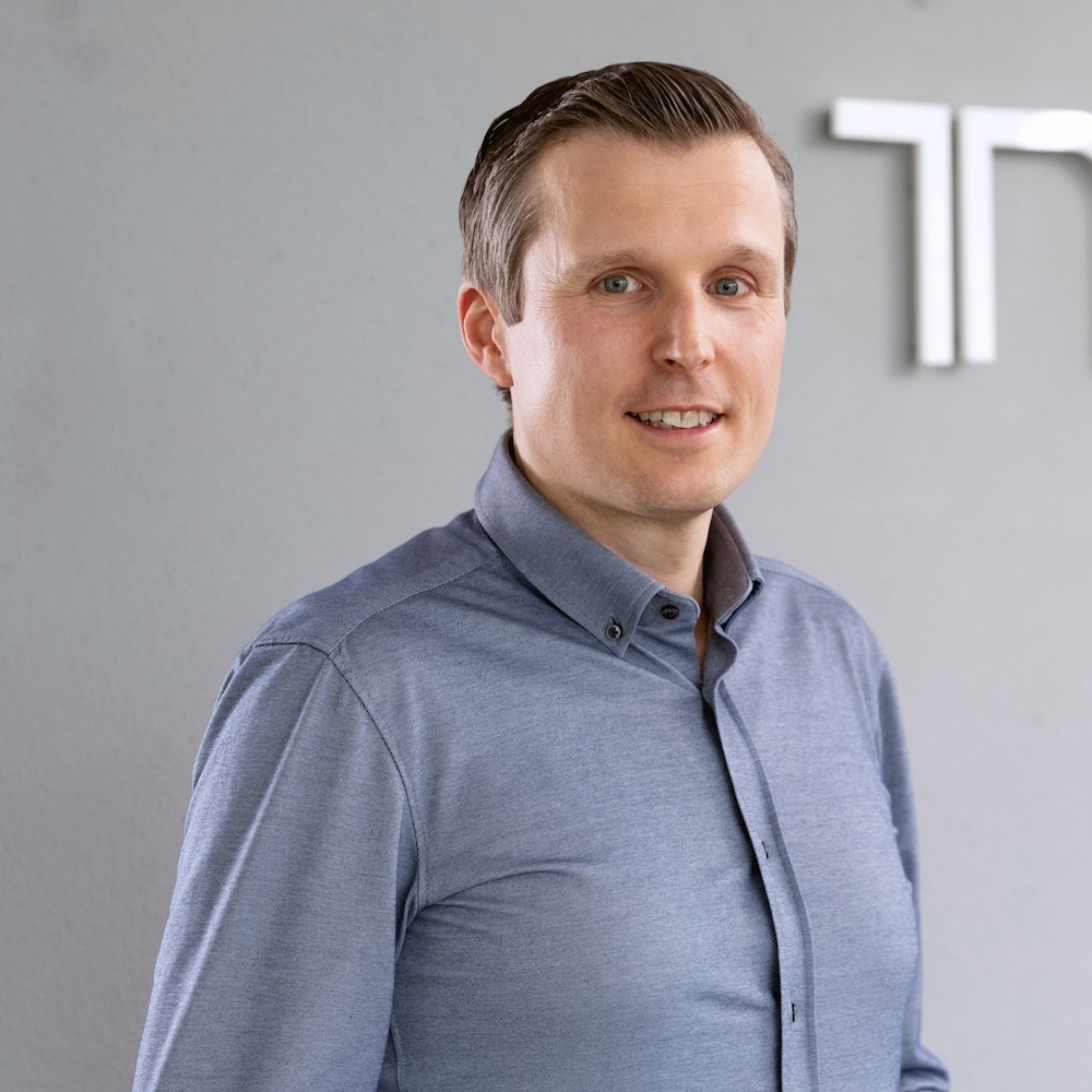 Dr. André Kortmann, der in einem hellblauen Button-up-Hemd vor einer grauen Wand steht.