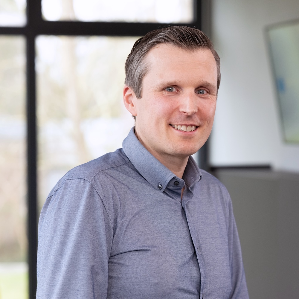 Dr. André Kortmann, der in einem hellblauen Button-up-Hemd in einer hellen, modernen Büroumgebung steht, mit großen Fenstern im Hintergrund.