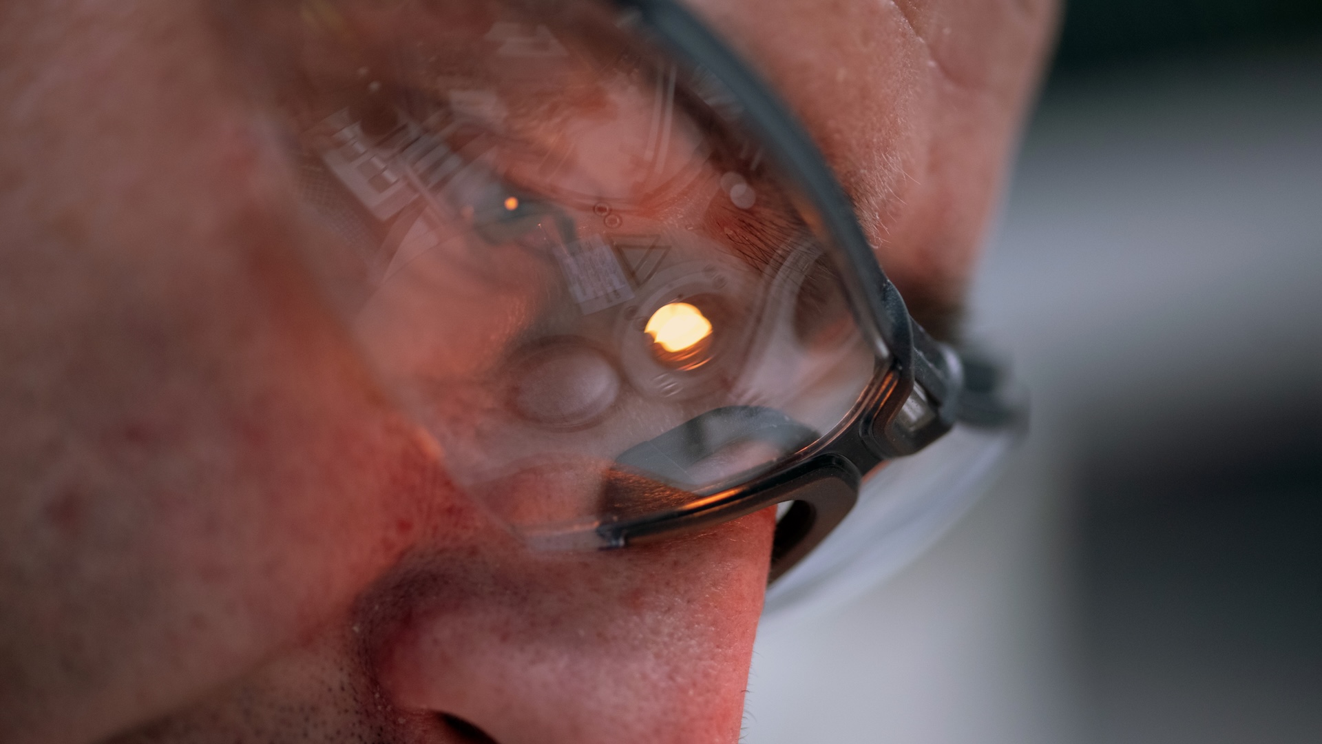 Nahaufnahme einer Person, die eine Schutzbrille trägt, mit – in den Brillengläsern sichtbar – der Reflexion eines optischen Geräts von ZEISS und einem leuchtenden Licht.