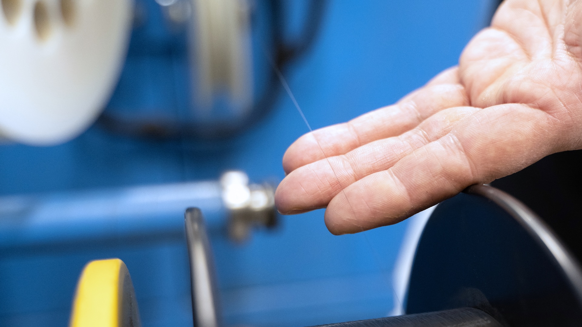 Nahaufnahme einer Hand, die einen feinen Glasfaserstrang inspiziert, der auf einer Präzisionsmaschine in einer ZEISS Fertigungsumgebung bearbeitet wird.