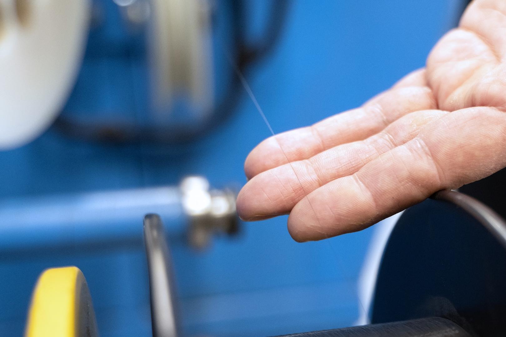 Nahaufnahme einer Hand, die einen feinen Glasfaserstrang inspiziert, der auf einer Präzisionsmaschine in einer ZEISS Fertigungsumgebung bearbeitet wird.