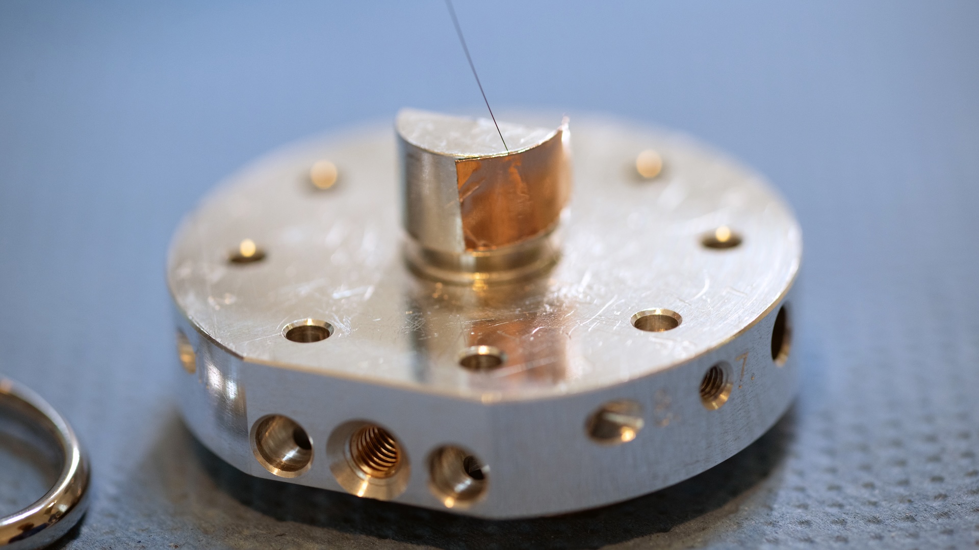Nahaufnahme eines präzisionsgefertigten Metallteils mit mehreren Bohrungen und einem dünnen Draht auf einer strukturierten Oberfläche für ZEISS.