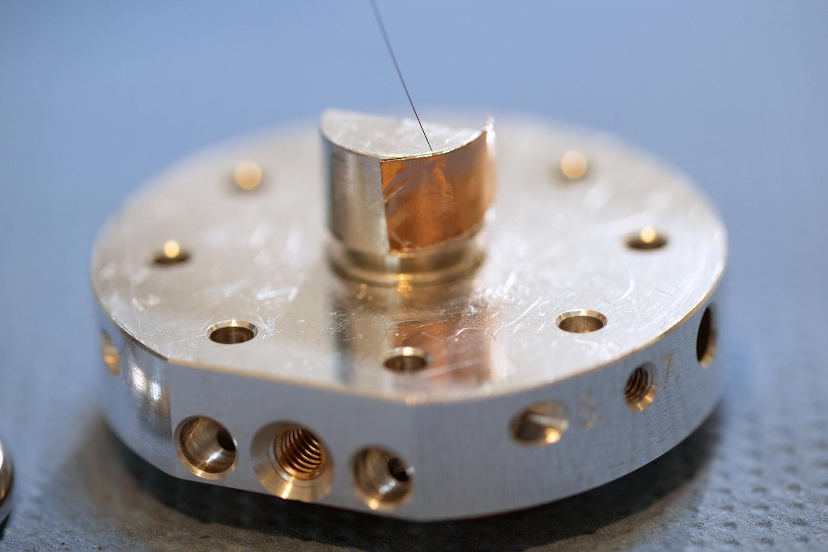 Nahaufnahme eines präzisionsgefertigten Metallteils mit mehreren Bohrungen und einem dünnen Draht auf einer strukturierten Oberfläche für ZEISS.