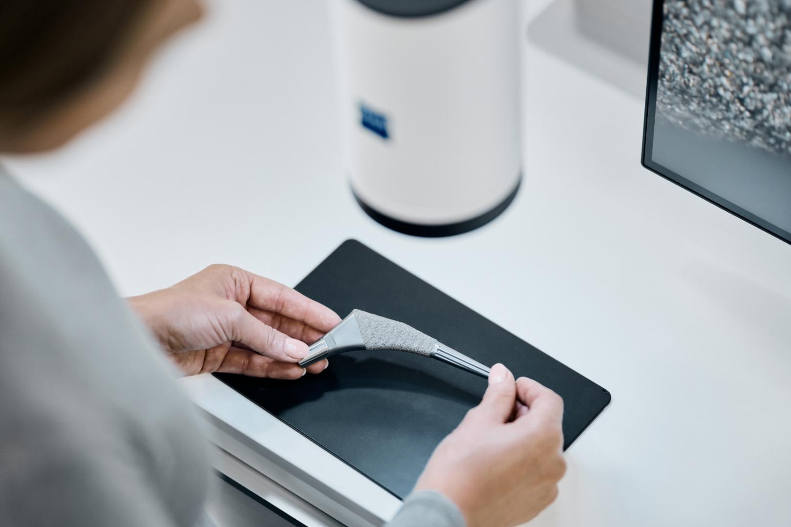 Eine Person hält ein 3D-gedrucktes Teil auf einer schwarzen Fläche neben einem ZEISS Gerät und einem Monitor, der eine vergrößerte Materialstruktur anzeigt.