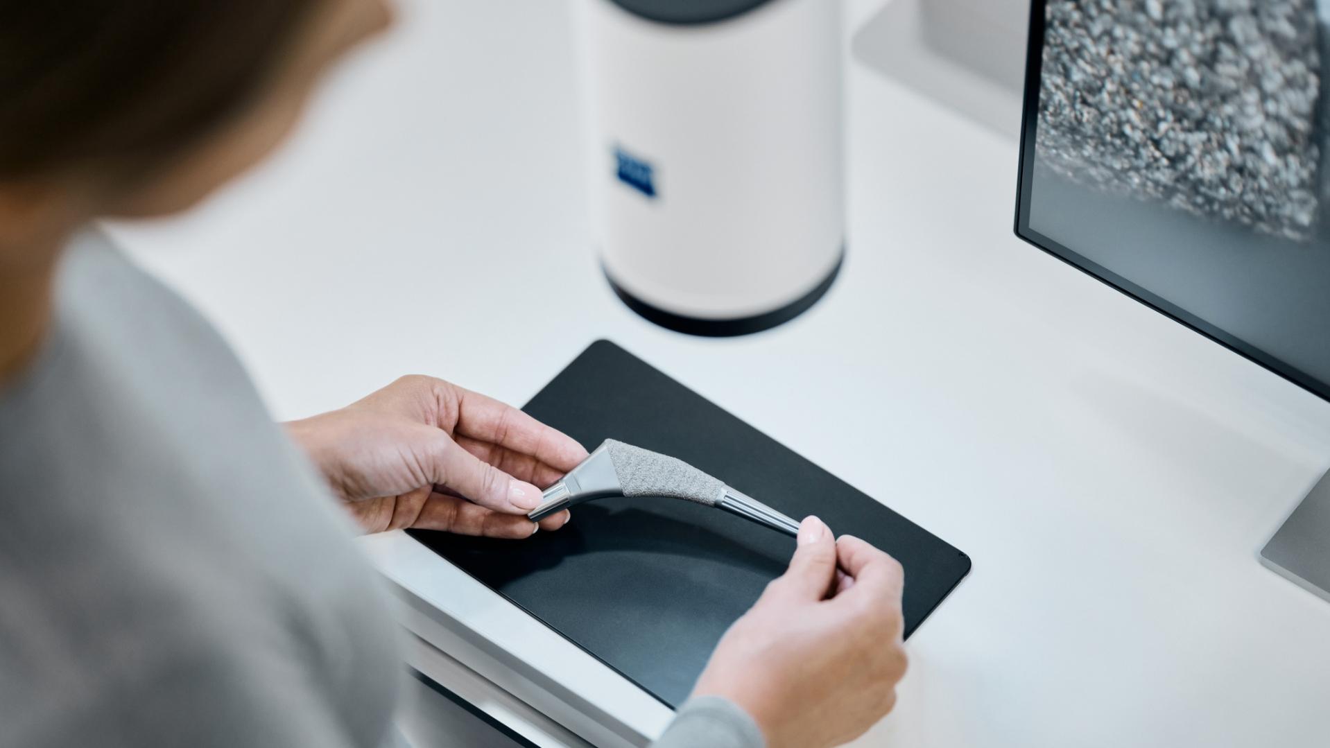 Eine Person hält ein 3D-gedrucktes Teil auf einer schwarzen Fläche neben einem ZEISS Gerät und einem Monitor, der eine vergrößerte Materialstruktur anzeigt.