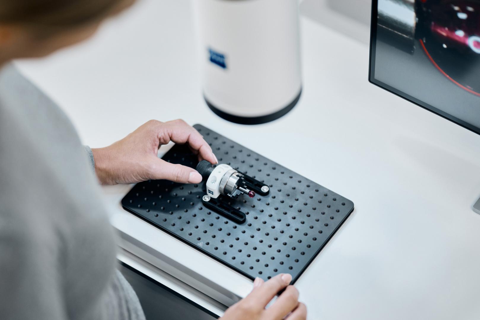 Eine Person positioniert ein mechanisches Bauteil auf einer perforierten Grundplatte neben einem ZEISS Gerät und einem Computermonitor, der eine Messschnittstelle anzeigt.