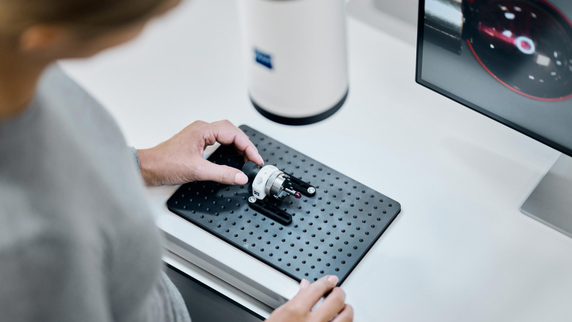 Eine Person positioniert ein mechanisches Bauteil auf einer perforierten Grundplatte neben einem ZEISS Gerät und einem Computermonitor, der eine Messschnittstelle anzeigt.