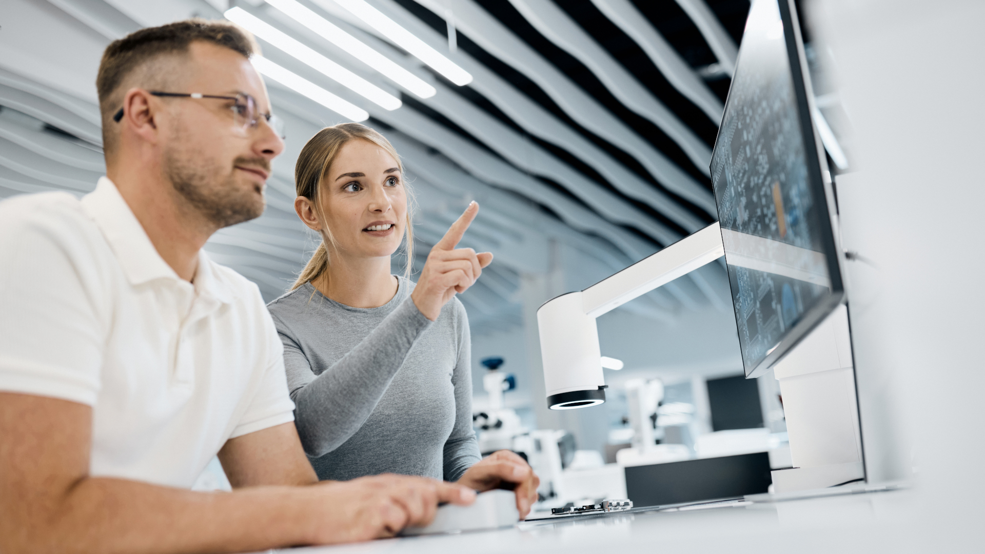 Zwei Fachkräfte arbeiten in einem modernen Labor zusammen und analysieren Daten auf einem großen Monitor, der an das ZEISS Smartzoom 100 angeschlossen ist.