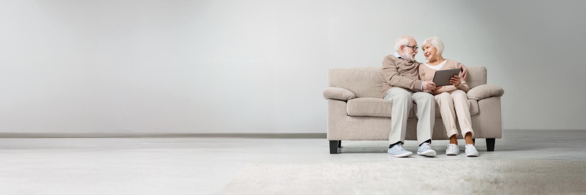 Ein älteres Paar sitzt eng zusammen auf einem beigen Sofa und liest ein Buch vor einem schlichten Hintergrund zu sehen ist.