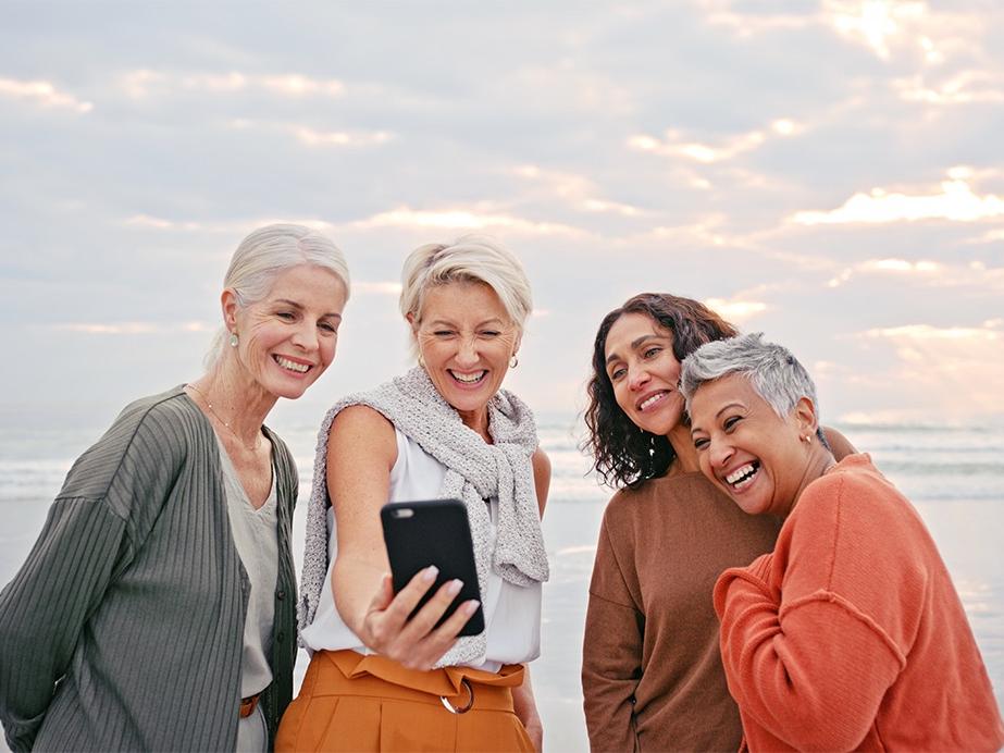 Vier lächelnde Frauen machen gemeinsam ein Selfie an einem Strand bei Sonnenuntergang.