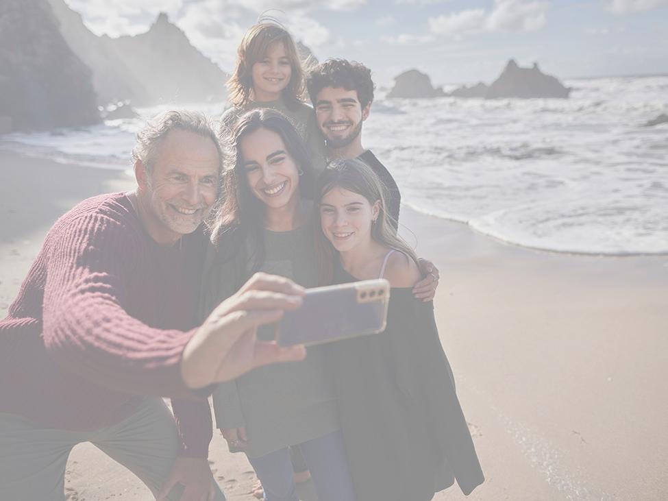Eine Gruppe von fünf Personen lächelt und macht gemeinsam ein Selfie an einem Strand mit Wellen und felsigen Klippen im Hintergrund.