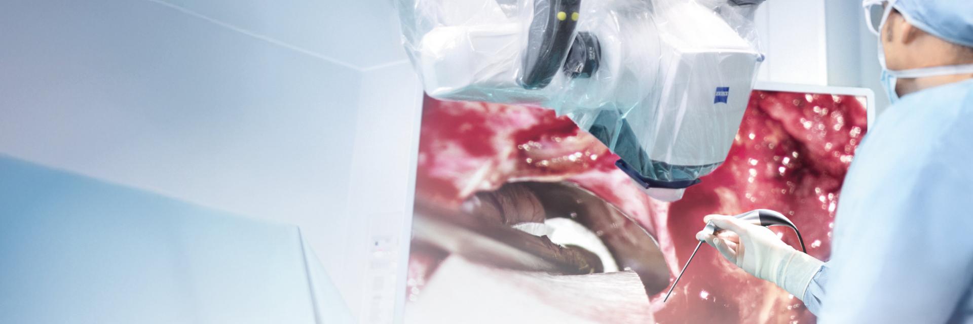 A medical professional operates using a ZEISS surgical microscope while viewing a high-resolution image of the procedure on a monitor.