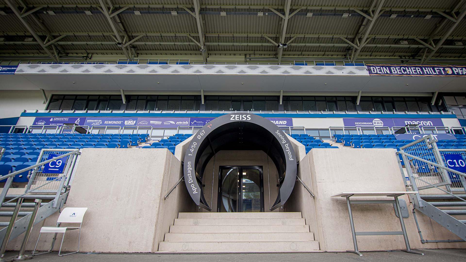 Blick auf den Eingangstunnel des Jenaer Fußballstadions mit „ZEISS“-Branding, umgeben von blauen Sitzen und Betonstufen unter einer überdachten Tribüne.