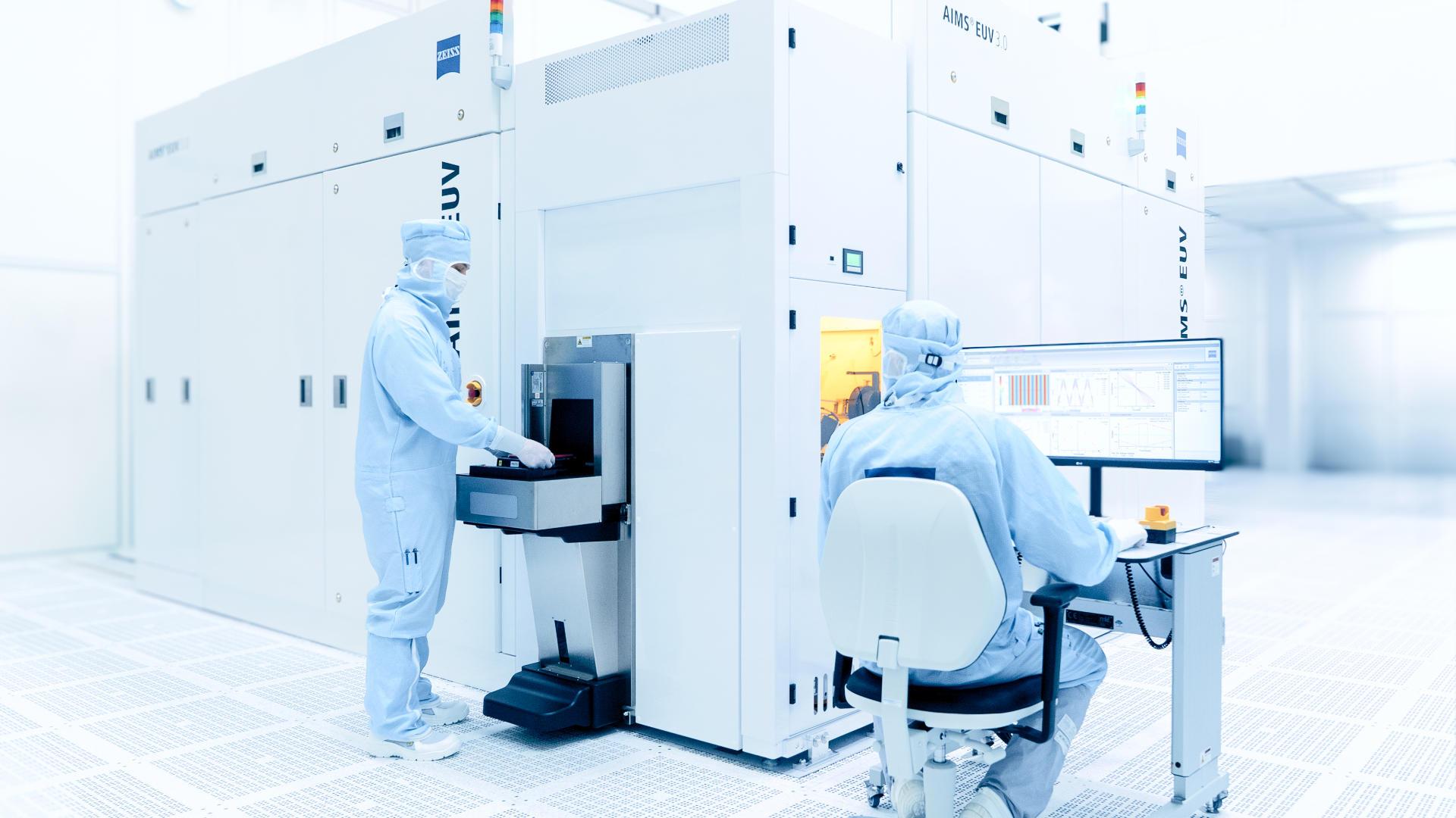 In einem weißen und hell erleuchteten Reinraum steht ein Gerät zur Maskenqualifizierung für die Halbleiterproduktion. Zwei Menschen in Schutzanzügen arbeiten am Gerät.