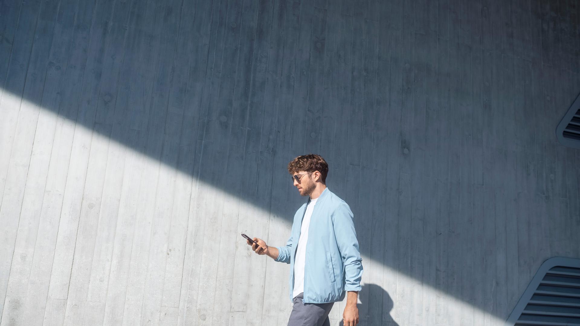 Ein Mann mit Sonnenbrille im Gesicht und Smartphone in der Hand steht seitlich vor grauer Betonwand., die schräg unten in der Sonne, schräg oben im Schatten ist.