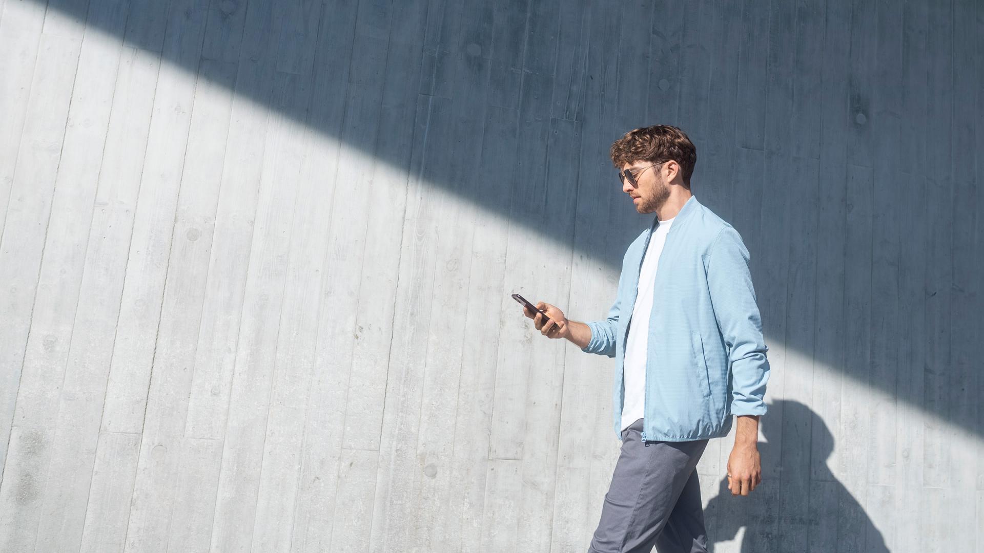 Ein Mann mit Sonnenbrille im Gesicht und Smartphone in der Hand steht seitlich vor grauer Betonwand., die schräg unten in der Sonne, schräg oben im Schatten ist.