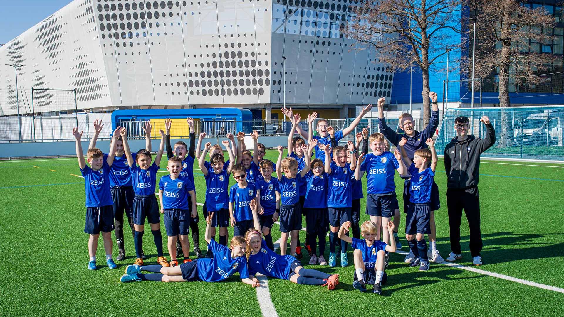 Willy Engelbrecht, Leiter der FCC-Fußballschule (links) und Patrick Paul, ZEISS CSR-Team (rechts) mit 23 Kindern von ZEISS Mitarbeitenden beim ersten Spieltagscamp am 4. April 2025.
