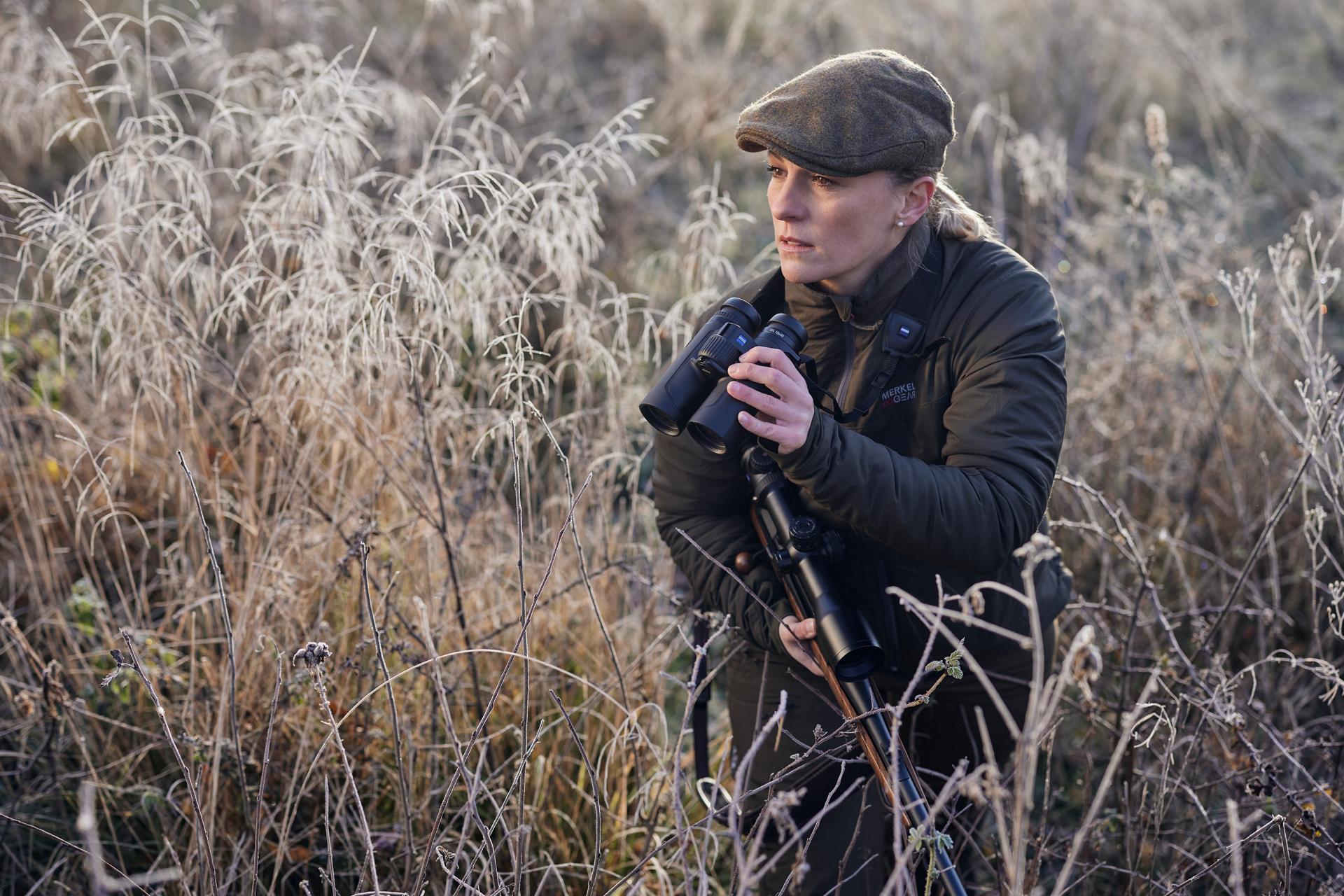 Eine Person in Outdoor-Kleidung hält ein Fernglas von ZEISS und ein Gewehr in der Hand, während sie auf einem frostigen Feld steht, das von hohem Gras umgeben ist.