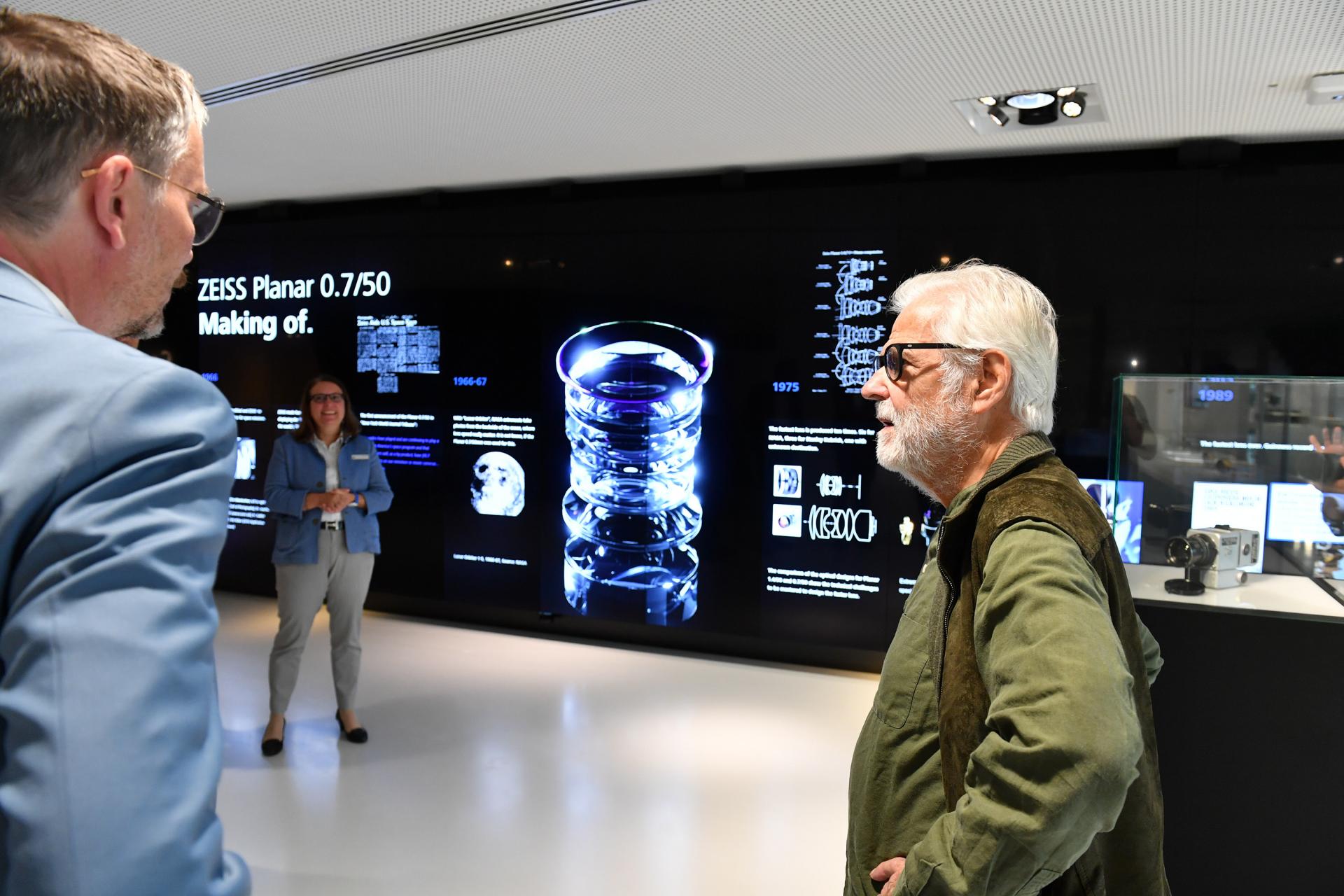 Drei Personen stehen vor einem ZEISS-Display, auf dem die Herstellung des ZEISS Planar 0,7/50-Objektivs mit technischen Abbildungen und Produktinformationen gezeigt wird, und unterhalten sich.