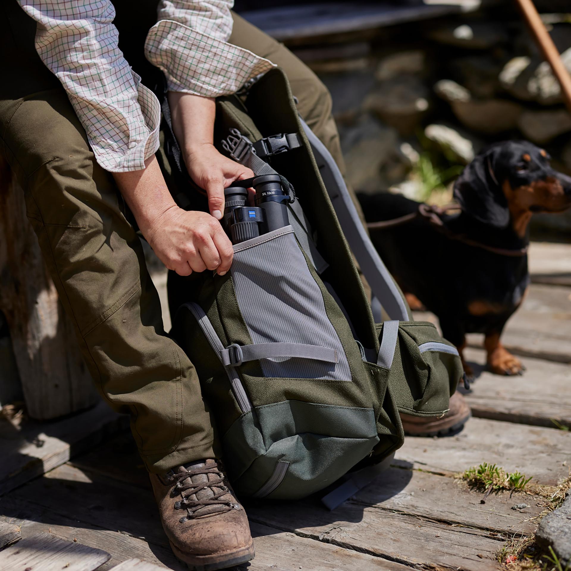 Eine Person steckt ein Fernglas von ZEISS in die Seitentasche eines grünen Rucksacks, während sie draußen neben einem kleinen schwarz-braunen Hund sitzt.