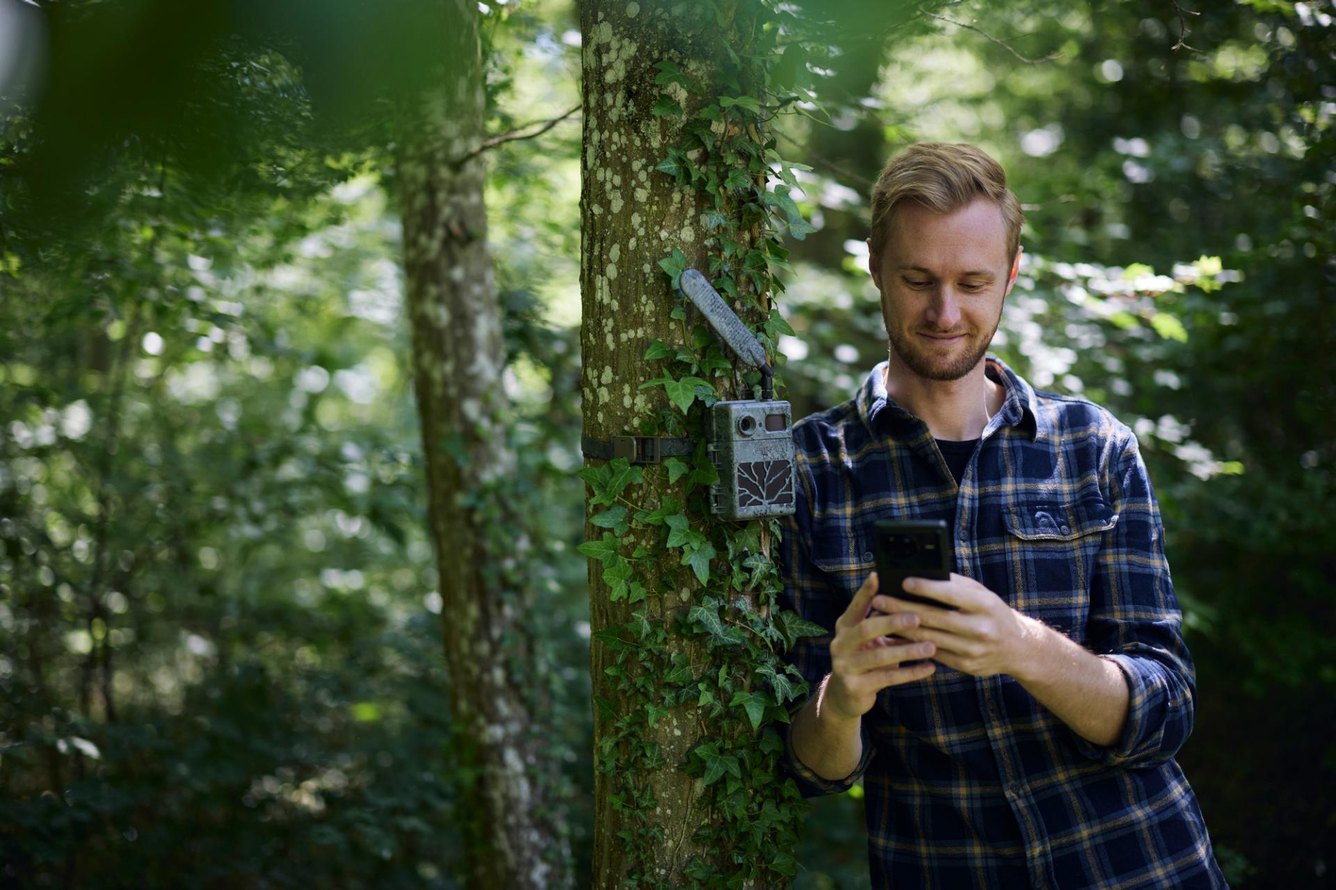Ein Mann steht in einem Wald und schaut auf sein Smartphone neben einer Wildkamera, die an einem Baum angebracht ist. Dabei ist Ausrüstung von ZEISS zu sehen.