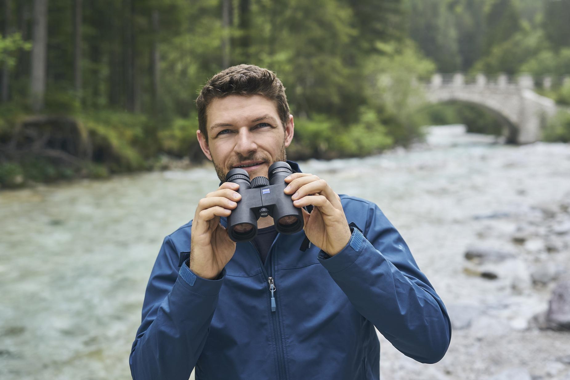 Ein Mann in einer blauen Jacke hält ein Fernglas von ZEISS in der Hand, während er neben einem Fluss in einem Waldgebiet steht, im Hintergrund ist eine Steinbrücke zu sehen.
