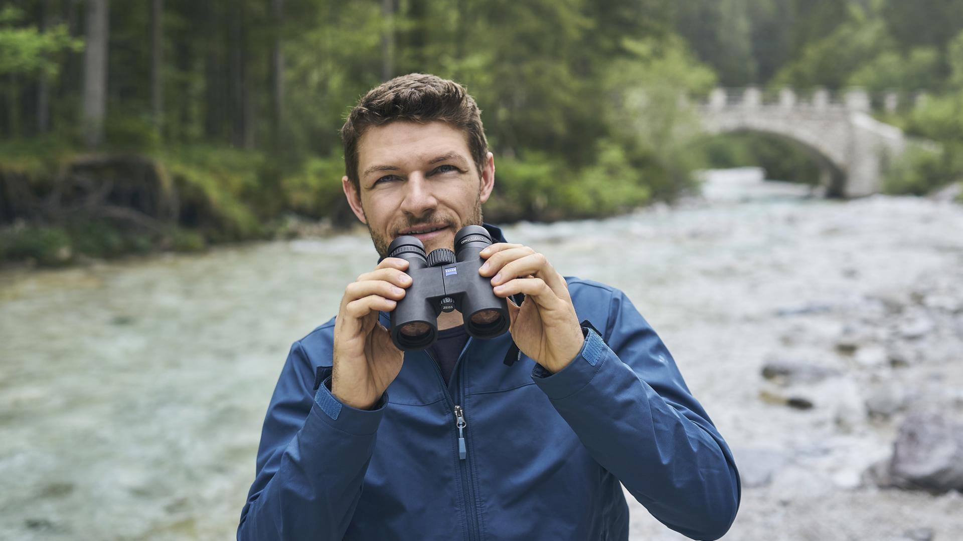 Ein Mann in einer blauen Jacke hält ein Fernglas von ZEISS in der Hand, während er neben einem Fluss in einem Waldgebiet steht, im Hintergrund ist eine Steinbrücke zu sehen.