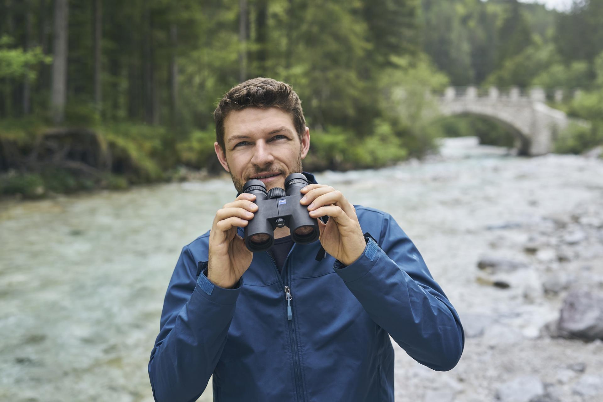 Zwei Mäner stehen vor einer Holzhütte im Wald, einer mit einem Fernglas, einer mit einem Jagdgewer in der Hand.