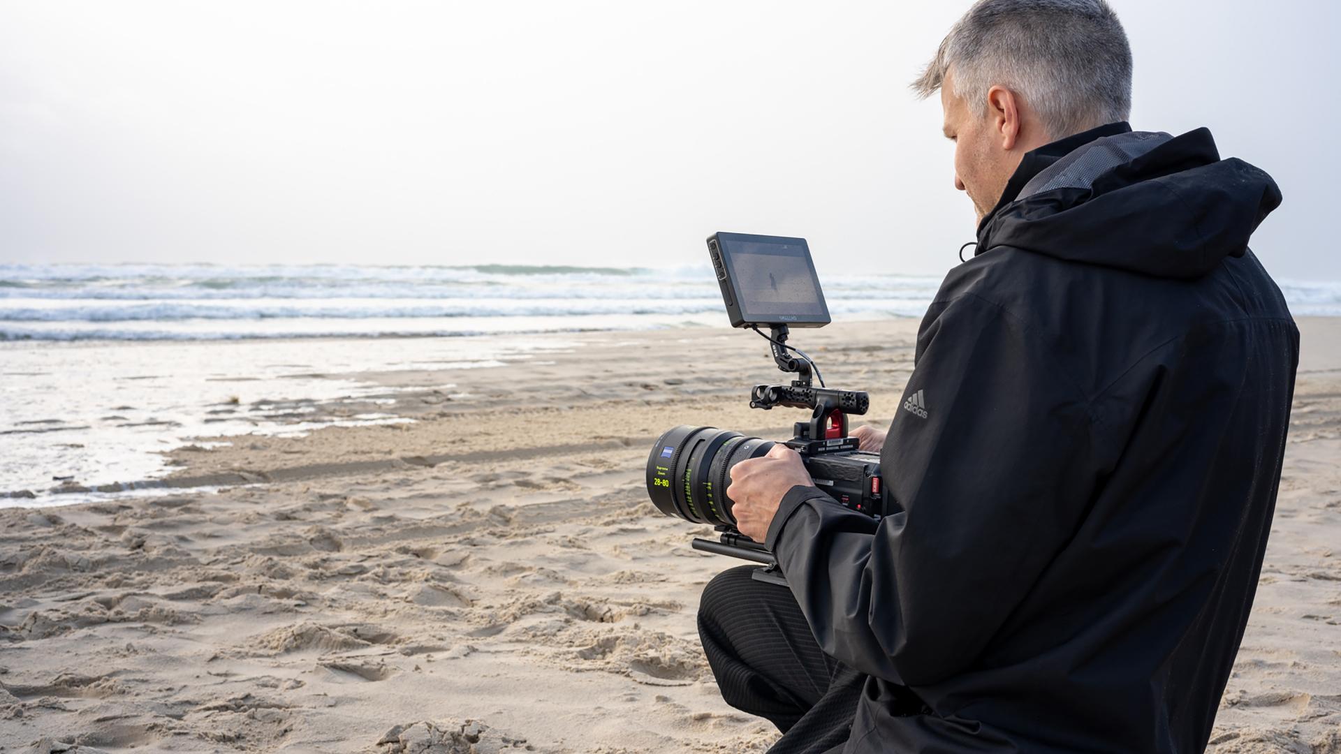 Ein Mann kniet an einem Sandstrand und bedient eine professionelle Videokamera, die mit einem ZEISS-Objektiv ausgestattet ist.