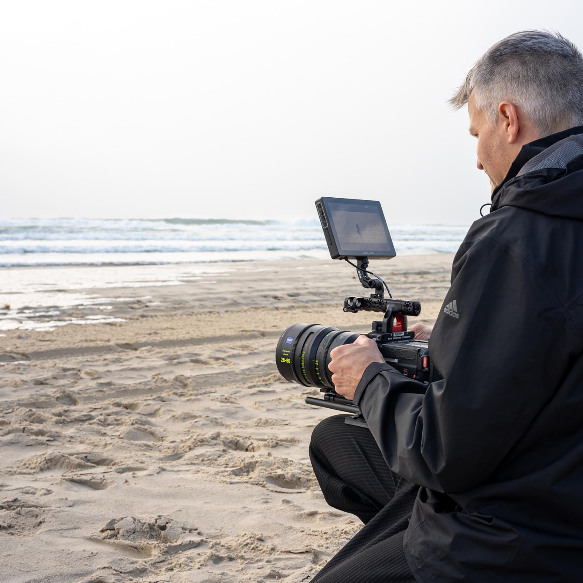 Ein Mann kniet an einem Sandstrand und bedient eine professionelle Videokamera, die mit einem ZEISS-Objektiv ausgestattet ist.