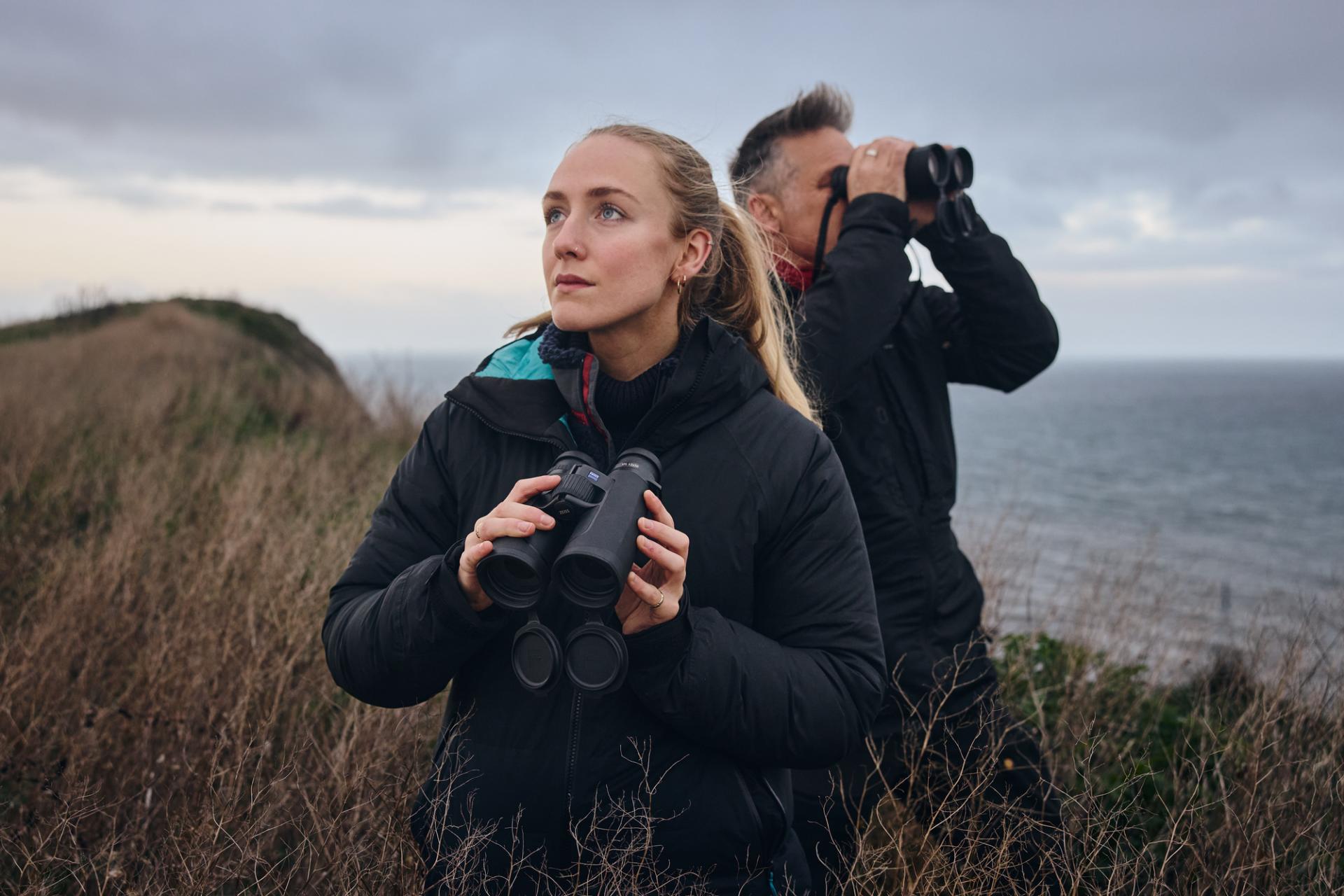 Zwei Personen stehen auf einer Küstenklippe und beobachten mit einem Fernglas von ZEISS die Landschaft.