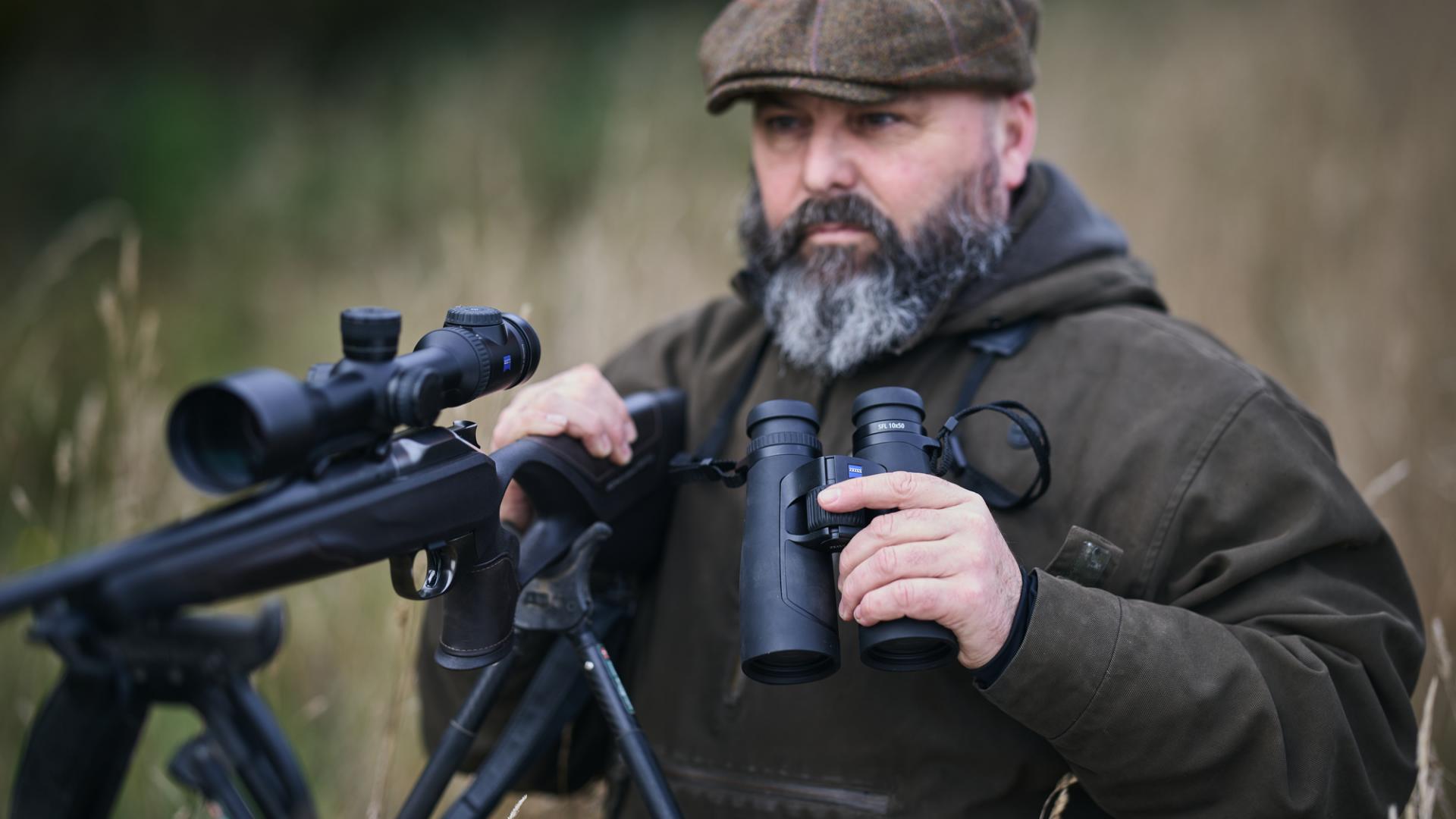 Ein bärtiger Mann in Outdoor-Kleidung steht auf einem Feld und hält ein Fernglas von ZEISS und ein Gewehr mit einem Zielfernrohr von ZEISS in den Händen.