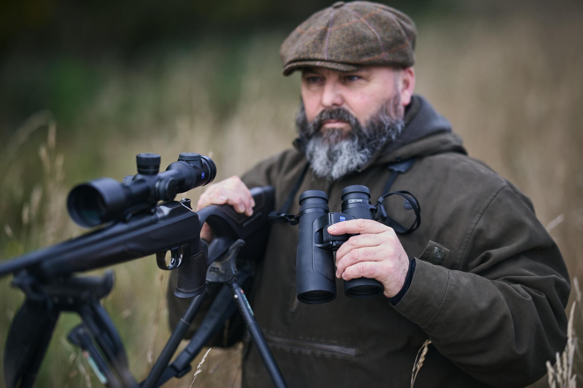 Ein bärtiger Mann in Outdoor-Kleidung steht auf einem Feld und hält ein Fernglas von ZEISS und ein Gewehr mit einem Zielfernrohr von ZEISS in den Händen.
