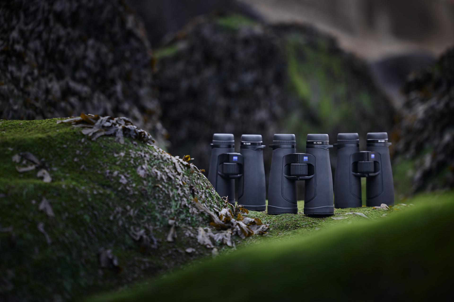 Eine Reihe schwarzer ZEISS Ferngläser ist im Freien auf moosbewachsenen Felsen ausgestellt.
