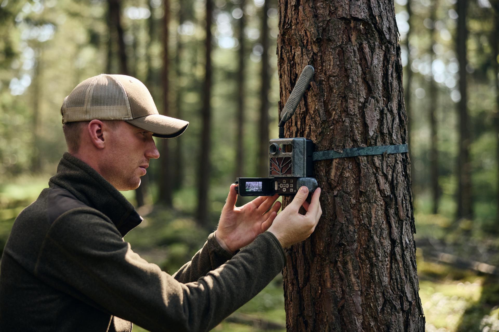 Ein Mann in Outdoor-Kleidung befestigt eine ZEISS-Wildkamera an einem Baum im Wald.