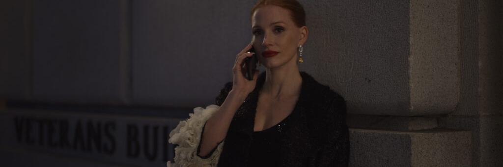 A woman in formal attire talks on a phone while standing against a stone wall near a sign that reads "VETERANS BUI" in dim evening lighting, showcasing the low-light performance of a ZEISS lens.