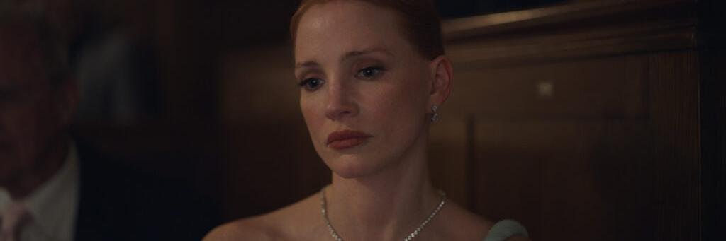 A woman with red hair wearing a formal dress and diamond necklace sits in a dimly lit audience, looking somber.