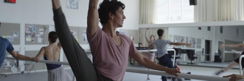 A group of ballet dancers practice at a barre in a brightly lit studio, with one dancer in the foreground performing a high leg extension.